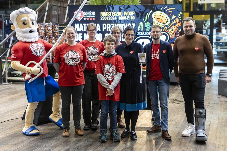 Kilka osób, większość w czerwonych koszulkach, stoi obok siebie. Po lewej w twarzy Mikołaja, w środku kobieta w okularach (prezydent Gdańska)