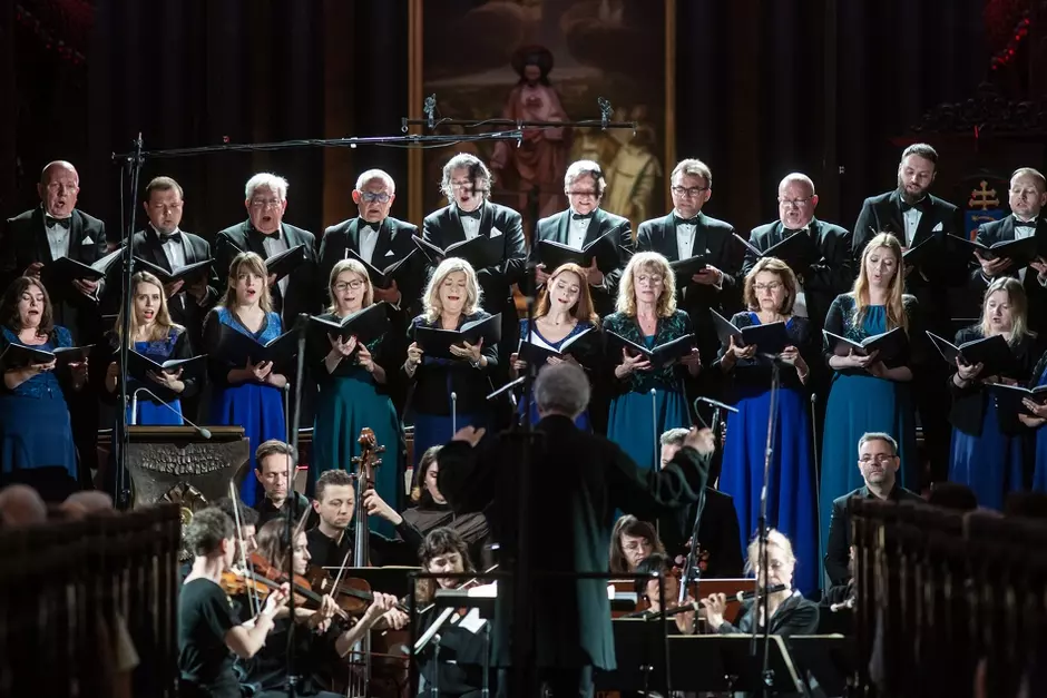 20 chórzystów stoi na scenie w dwóch rzędach, z tyłu mężczyźni, z przodu kobiety, przed nimi siedzą muzycy, na samym przedzie dyrygent