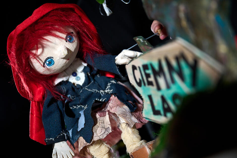 lalka przedstawiająca dziewczynkę z rudymi włosami przykrytymi częściowo czerwonym kapturem, obok niej tabliczka z napisem ciemny las