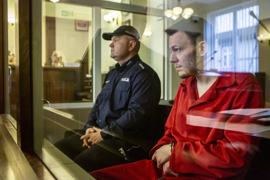 Na zdjęciu widać dwie osoby siedzące obok siebie w pomieszczeniu, które wygląda na salę sądową. Po lewej stronie jest osoba w policyjnym mundurze z napisem POLICJA  na rękawie, co wskazuje na to, że może to być policjant. Osoba ta ma krótkie ciemne włosy i patrzy przed siebie z poważnym wyrazem twarzy. Po prawej stronie siedzi mężczyzna w czerwonym ubraniu, które może być strójem więziennym lub innym rodzajem jednolitego ubioru. Mężczyzna ma krótkie ciemne włosy, brodę i patrzy w bok z zamyśloną miną. Obie postacie znajdują się za coś w rodzaju przezroczystej przegrody, być może jest to zabezpieczenie lub bariera ochronna.