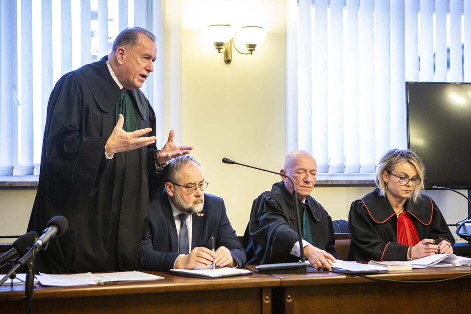 Na zdjęciu widoczna jest grupa osób w sali sądowej. Po lewej stronie stoi mężczyzna w togach sędziowskich, wydaje się aktywnie gestykulować i mówić, co sugeruje, że może przemawiać lub argumentować sprawę. Jego wyraz twarzy wydaje się dość ekspresyjny. Po prawej stronie mężczyzny siedzą trzy osoby przy stole. Pierwsza od lewej to mężczyzna w okularach, który również ma na sobie togi sędziowskie i wydaje się koncentrować na pisaniu lub przeglądaniu dokumentów. Obok niego siedzi drugi mężczyzna w togach, który ma zamknięte oczy i wygląda na zamyślonego lub być może zmęczonego. Trzecia osoba to kobieta, także w togach sędziowskich z charakterystycznym czerwonym jabotem, skupiona na pisaniu lub czytaniu dokumentu przed nią.