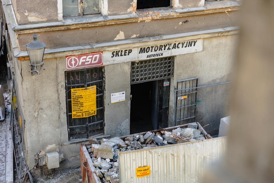 zdjęcie zniszczonej kamienicy o brązowej elewacji, widać też otwarte drzwi do lokalu na parterze i nad nim szyld z napisem salon motoryzacyjny