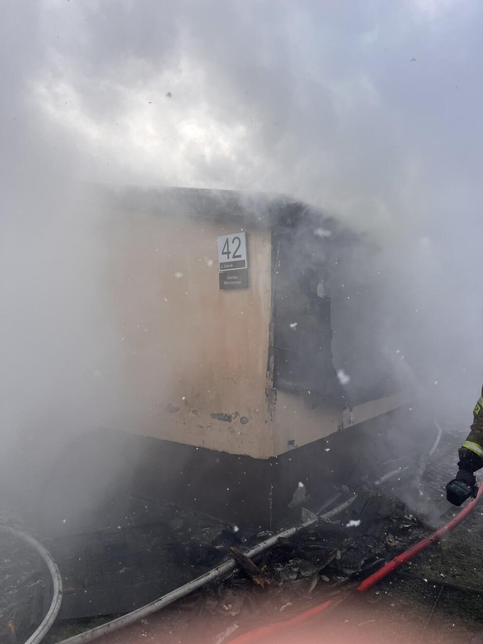 Z dymu wyłania się ściana domu z numerem przy drzwiach wejściowych.