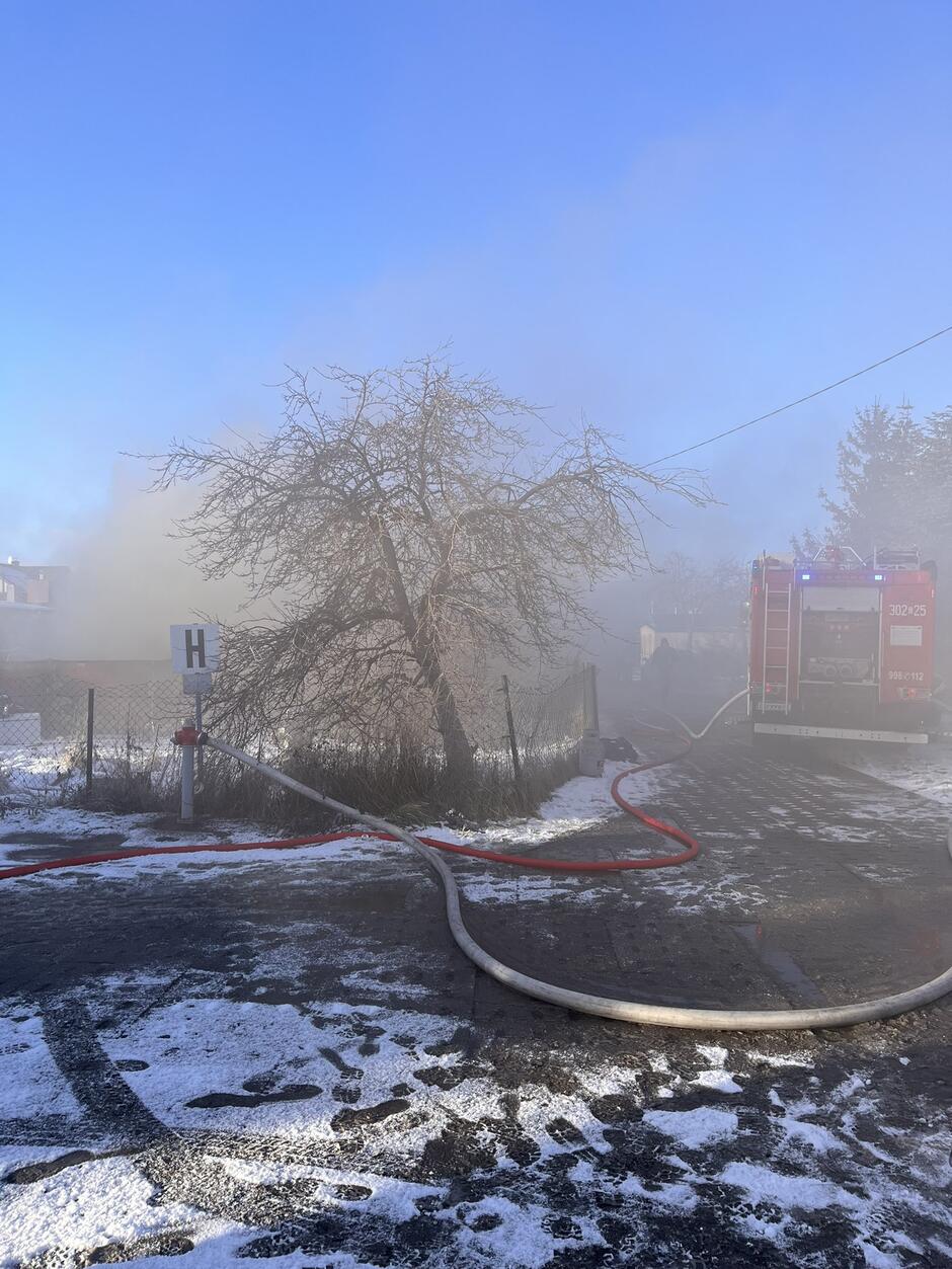 Na zdjęciu widać pochylone drzewo i dużo dymu. Po prawej stronie stoi czerwony strażacki wóz, na ziemi okrytej nieregularnie śniegiem leży wąż strażacki.