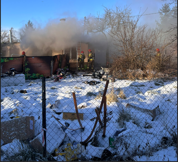 Na zdjęciu widać chmurę dymu, która spowija parterowy domek. U góry niebieskie niebo, obok domu drzewa bez liści, zimowe, przed domem plac pokryty śniegiem, najbliżej płot z siatki metalowej, pochylony.