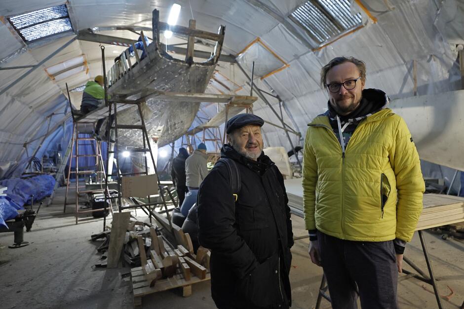 Dwaj mężczyźni w hali, na tle remontowanego jachtu - po lewej stoi niski starszy mężczyzna, po prawej młody, wysoki 