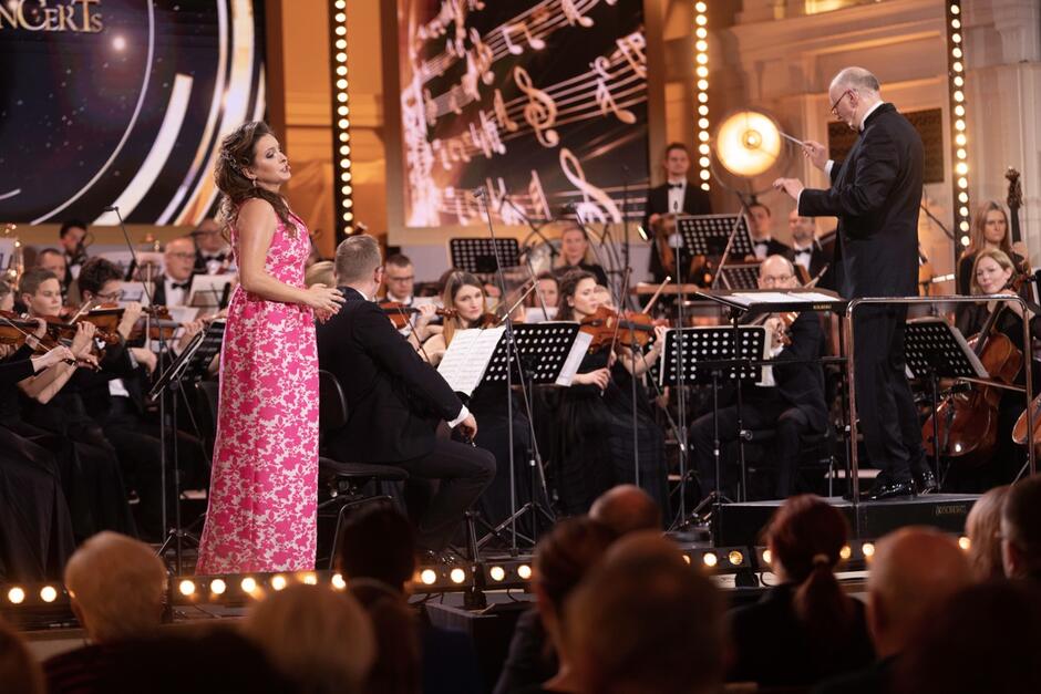 Solistka w różowej długiej sukni podczas koncertu, w tle siedzący muzycy, obok stojący dyrygent
