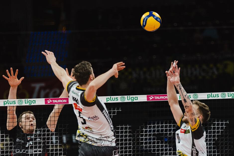 Dwóch siatkarzy w białych koszulkach w wyskoku: po lewej szykuje się do ataku, po prawej rozgrywał. Nad nimi piłka, przed nimi siatka