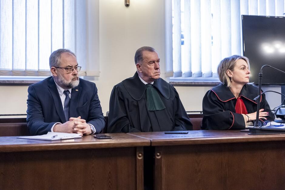 Na zdjęciu widać trzech ludzi siedzących obok siebie w pomieszczeniu, które wygląda na salę sądową. Osoba po lewej stronie to mężczyzna w średnim wieku, ubrany w ciemny garnitur, z brodą, okularami i odznaką na klapie, co może sugerować jego oficjalną rolę lub tytuł. Wydaje się, że ma złożone ręce i patrzy przed siebie, z poważną miną. W środku jest mężczyzna w starszym wieku, ubrany w czarną togę sędziowską z zieloną apliką, co wskazuje na jego zawód jako sędziego. Ma poważne i skupione wyraz twarzy, co może odzwierciedlać wagę spraw sądowych, w których uczestniczy. Osoba po prawej to kobieta w średnim wieku, ubrana w czarno-czerwoną togę, co może sugerować jej rolę jako sędziego lub adwokata. Jej włosy są krótkie i blond, a postawa zwrócona w stronę ekranu lub osoby poza kadr, co może świadczyć o uwadze, jaką poświęca na bieżące wydarzenia w sali.