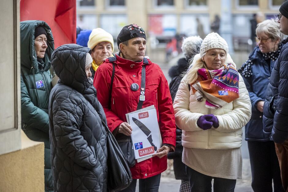 Zdjęcie przedstawia grupę osób w średnim i starszym wieku ubranych w zimowe kurtki i czapki, co wskazuje na chłodną pogodę. Skupiają się one na czymś poza kadr. W centrum obrazu widzimy mężczyznę w czerwonej kurtce trzymającego tabliczkę z napisem, co sugeruje, że uczestniczą w jakimś zgromadzeniu lub demonstracji. Wyraz twarzy osób jest poważny, co może oznaczać, że temat ich zgromadzenia jest ważny lub kontrowersyjny. Tło jest nieostre, ale można zauważyć miejską architekturę, co sugeruje, że wydarzenie ma miejsce na ulicy lub w przestrzeni publicznej.