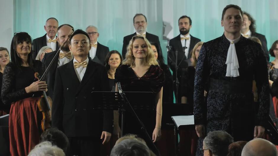 Trzy osoby elegancko ubrane, za nimi muzycy z instrumentami