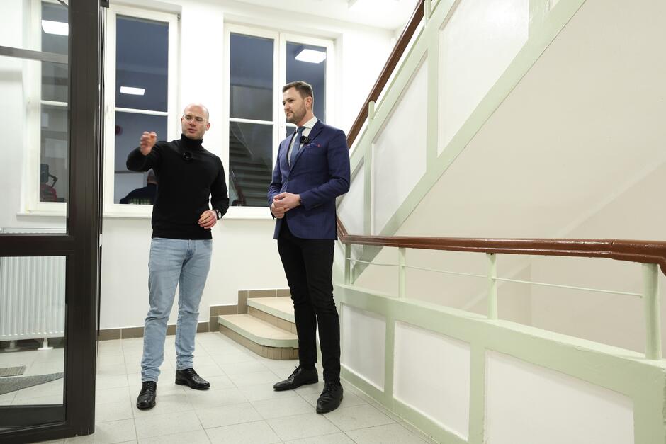 na zdjęciu dwóch szczupłych wysokich mężczyzn, stoją na klatce schodowej, mężczyzna po lewej wskazuje ręką na drzwi