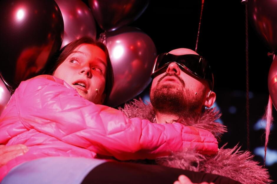 mężczyzna w futurystycznych okularach przeciwsłonecznych trzyma na rękach dziewczynę, oboje skąpani w czerwono-różowym świetle