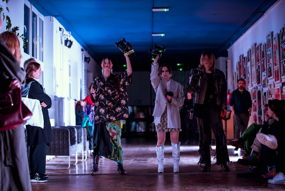 troje młodych performerów stoi na korytarzu, pokazuje coś publiczności