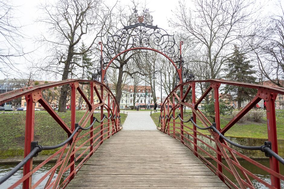 na zdjęciu mostek z metalowymi dekoracyjnymi balustradami w kolorze rdzawo - czerwonym, w tle drzewa