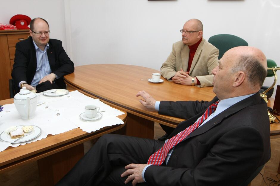 Trzej mężczyźni w średnim wieku siedzą wokół biurka i rozmawiają