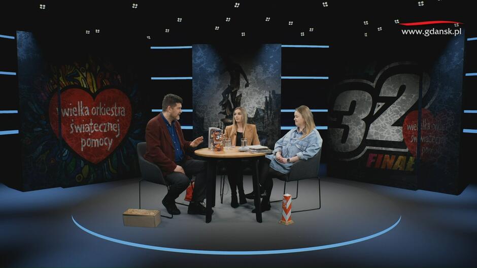 na zdjęciu studio telewizyjne, przy okrągłym stoliku siedzą na krzesłach dwie młode kobiety i jeden wysoki mężczyzna w średnim wieku