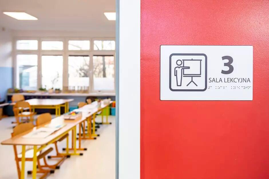 Po prawej stronie tablica z różnymi oznaczeniami i napisem: 3 sala lekcyjna. Po prawej przez otwarte drzwi widać krzesła i ławki szkolne