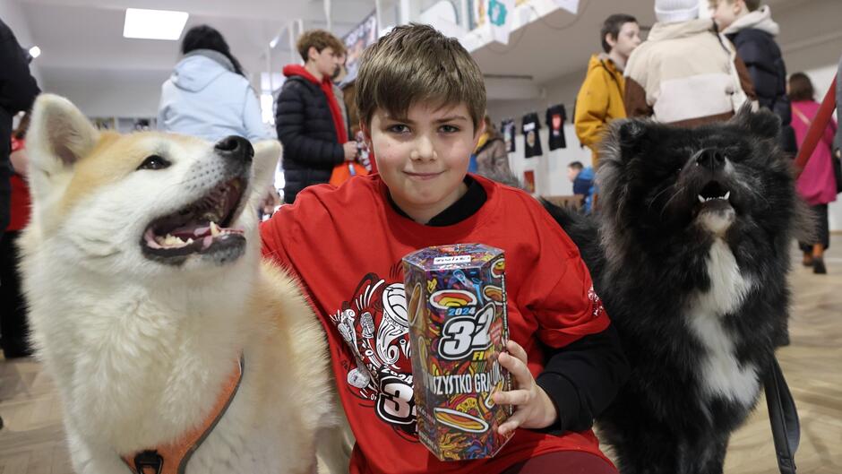 Na zdjęciu widzimy młodego chłopca siedzącego między dwoma psami. Chłopiec ma na sobie czerwoną koszulkę z grafiką i trzyma w rękach kolorowe pudełko z napisem 2024  i słowami  wszystko gra , co może sugerować jakiś element zabawki lub gry. Po jego prawej stronie znajduje się pies rasy akita inu, który ma otwartą pysk i wygląda na zadowolonego lub w trakcie wydawania dźwięku. Po lewej stronie chłopca jest ciemniejszy pies, który również ma otwartą pysk i wydaje się być w trakcie szczekania lub ziewania. Tło zdjęcia to wnętrze pomieszczenia, w którym znajduje się wiele osób, prawdopodobnie jest to jakaś publiczna przestrzeń lub wydarzenie z udziałem zwierząt. Atmosfera wydaje się być radosna i pełna życia