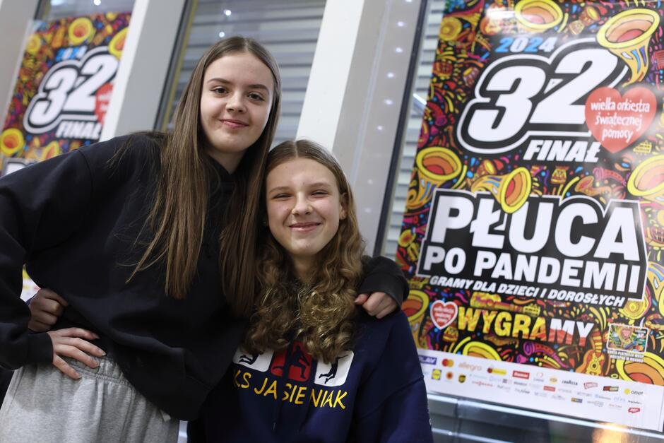Na zdjęciu widzimy dwie młode dziewczyny uśmiechające się do kamery. Stoją one przed kolorowym banerem z napisem 32 FINAŁ  i  PŁUCA PO PANDEMII . Tło sugeruje, że mogą uczestniczyć w wydarzeniu związanym z Wielką Orkiestrą Świątecznej Pomocy, polską organizacją charytatywną, która jest znana z organizacji corocznych finałów gromadzących środki na różne cele zdrowotne i społeczne. Dziewczyny mają na sobie ciemne bluzy, jedna z nich ma na bluzie napis, który może być związany z tym wydarzeniem. Wyrażają pozytywne emocje i wydają się zadowolone z uczestnictwa w tym wydarzeniu