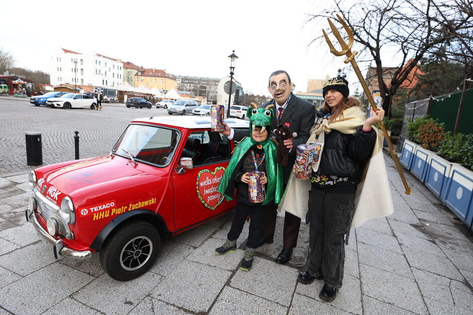 Mężczyzna w masce Jasia fasoli przed małym czerwonym samochodem z kobietą i dzieckiem, w dłoni trzyma puszkę WOŚP