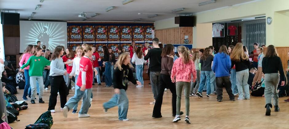 grupa kilkudziesięciorga uczniów, nastolatków tańczą na szkolnym holu, na ścianie plakaty WOŚP