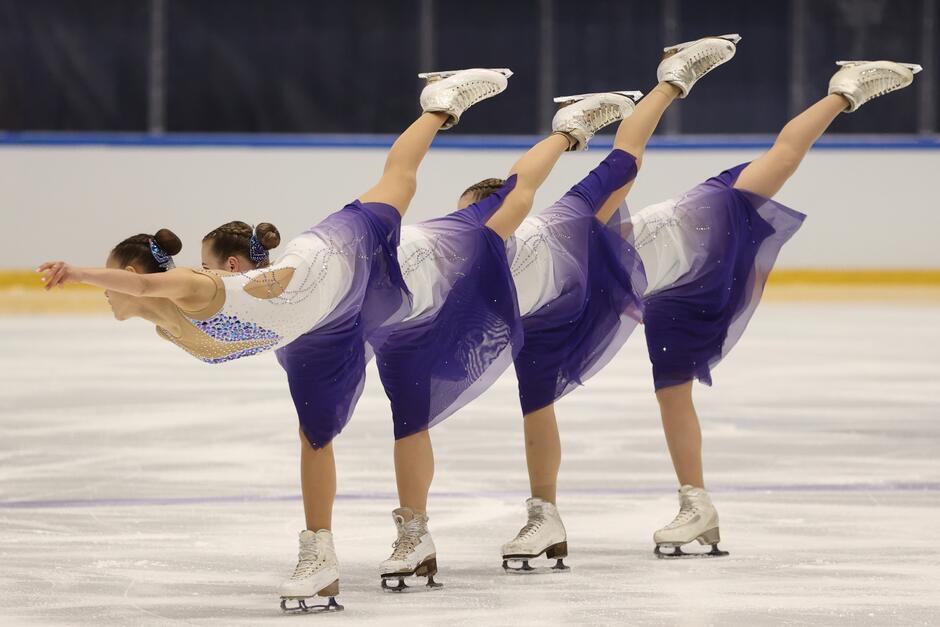 Trzy łyżwiarki jadą obok siebie, są pochylone do przodu, jedna noga wyprostowana do tyłu