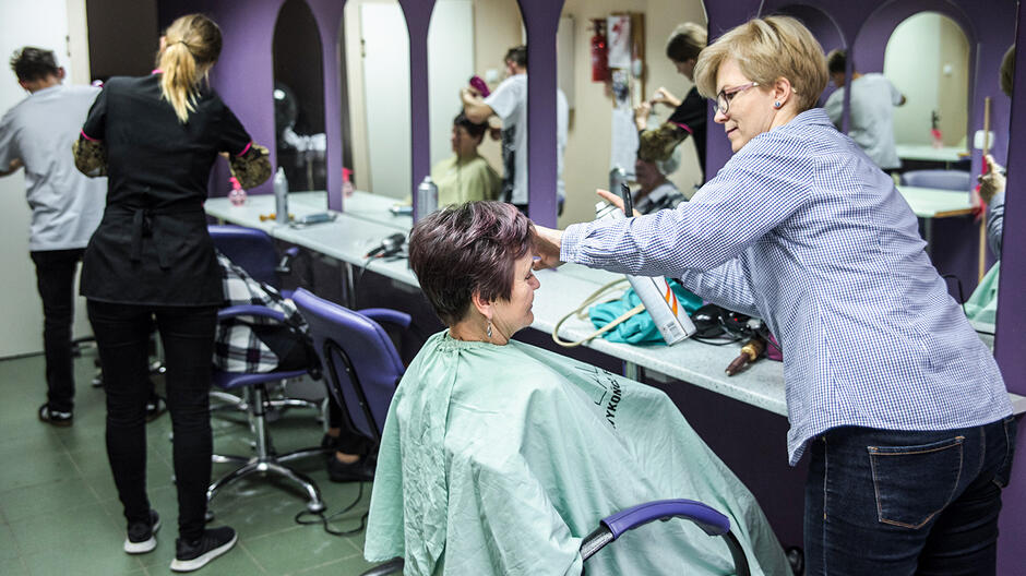 Wnętrze salonu fryzjerskiego, na pierwszym planie fryzjerka układa fryzurę starszej kobiecie siedzącej na fotelu, w tle widoczna druga fryzjerka z klientką na fotelu. Na ścianie rząd luster, w których odbija się wnętrze salonu, z klientami czekającymi na swoją kolej