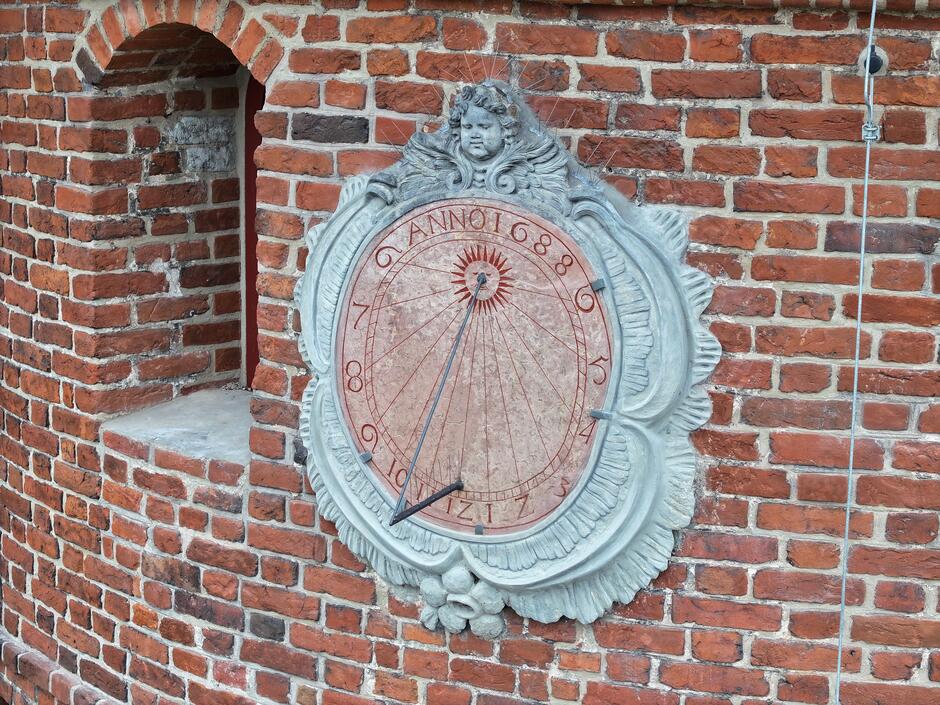 Na zdjęciu widzimy detal architektoniczny umieszczony na ceglanej ścianie – jest to kamienny słoneczny zegar. Zegar ma wyryte i malowane na czerwono rzymskie cyfry oraz linie wskazujące godziny, a w jego centralnej części znajduje się gnomon, czyli element wystający, który rzuca cień na tarczę zegara, umożliwiając odczytanie czasu słonecznego. Nad tarczą zegara umieszczona jest dekoracyjna płaskorzeźba przedstawiająca twarz, prawdopodobnie symbolizującą słońce, z promienistą aureolą wokół głowy i zdobieniami w formie liści na dolnej krawędzi. Na tarczy zegara widoczny jest napis ANNO 1686 , co wskazuje na rok, w którym zegar mógł być wykonany lub zamontowany. Cegły w tle mają różne odcienie czerwieni i są zróżnicowane pod względem kształtu i tekstury, co dodaje charakteru i wskazuje na historyczny charakter konstrukcji