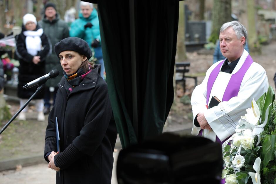 Zdjęcie przedstawia scenę z ceremonii pogrzebowej. Na pierwszym planie po lewej stronie widoczna jest kobieta w czarnym płaszczu i czapce, stojąca przed mikrofonem, najwyraźniej przygotowująca się do przemówienia lub właśnie je wygłaszająca. W ręce trzyma kartkę, co może sugerować, że jest to jej mowa lub notatki. Na twarzy ma poważny wyraz, co jest adekwatne do okoliczności. Po prawej stronie zdjęcia, w tle, stoi mężczyzna ubrany w liturgiczne szaty w kolorach fioletu i białego, typowe dla księży katolickich podczas okresu Wielkiego Postu lub pogrzebu. Jego ręce są złożone, a na twarzy ma poważny wyraz, co pasuje do roli duchownego przewodniczącego ceremonii pogrzebowej. W głębi za nimi widać grupę ludzi ubranych w ciepłe zimowe ubrania, co może sugerować, że pogrzeb odbywa się w chłodniejszym klimacie lub porze roku. Sceneria z dużą ilością drzew i nagrobków na cmentarzu dodaje do atmosfery żałobnego nastroju.