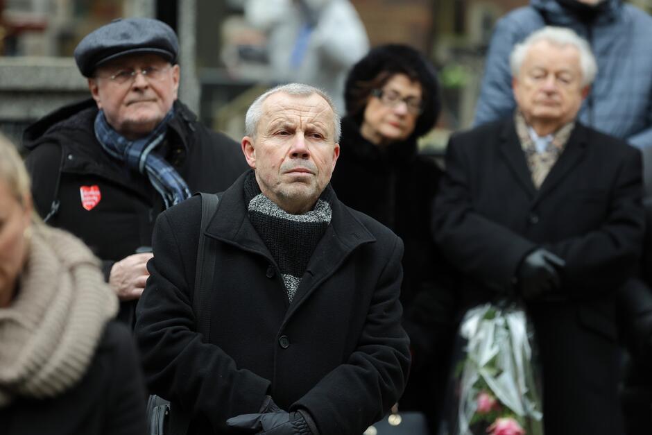 Zdjęcie pokazuje grupę ludzi, prawdopodobnie uczestników ceremonii pogrzebowej. Na pierwszym planie znajduje się mężczyzna o gołej głowie i wyrazie twarzy pełnym refleksji lub smutku. Jest ubrany w ciemny płaszcz i szalik, co może wskazywać na zimową porę. Za nim w tle są inne osoby. Mężczyzna po lewej stronie nosi ciemną kurtkę i czapkę oraz szalik, a na piersi ma przypięty czerwony symbol, który może być znakiem solidarności lub przynależności do jakiejś grupy. Kobieta w środku zdjęcia, również w ciemnym ubiorze, ma zamknięte oczy i wygląda na zamyśloną. Mężczyzna po prawej stronie, także ubrany na czarno, stoi z rękami skrzyżowanymi i ma zamknięte oczy. Wszystkie osoby na zdjęciu mają poważny wyraz twarzy i ubrane są w ciemne, eleganckie ubrania, co jest typowe dla uczestników pogrzebu. Ogólna atmosfera zdjęcia jest spokojna i pełna powagi.