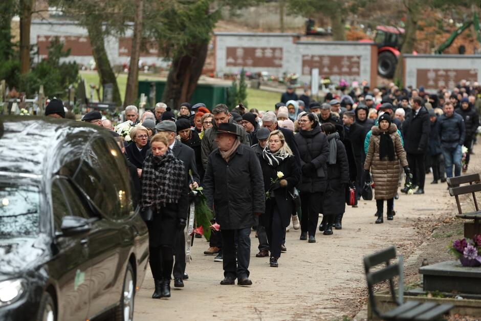 Na zdjęciu widoczny jest długi kondukt żałobny na cmentarzu. Przemarsz ludzi, którzy są ubrani w ciemne, formalne ubrania, wskazuje, że biorą oni udział w pogrzebie. Niektórzy z nich trzymają w rękach białe kwiaty. Grupa podąża za czarnym karawanem pogrzebowym, który jest częściowo widoczny na lewej stronie zdjęcia. Osoby w kondukcie mają poważne i smutne wyrazy twarzy, co jest typowe dla tego rodzaju uroczystości. W tle zdjęcia widać nagrobki i drzewa, co dodatkowo podkreśla kontekst cmentarny.