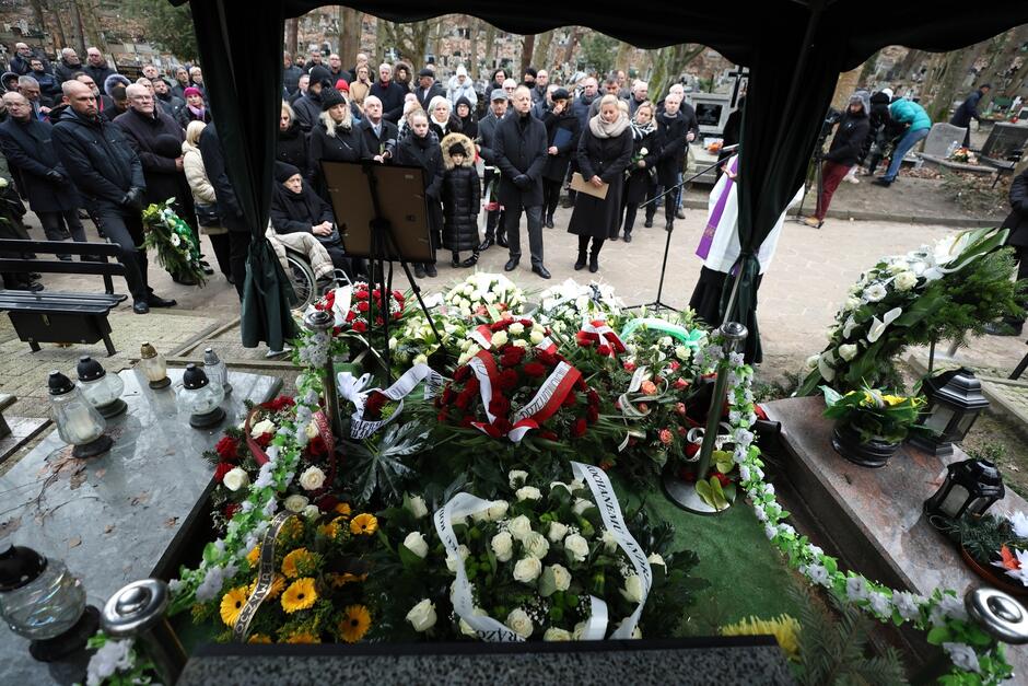 To zdjęcie przedstawia moment ceremonii pogrzebowej na cmentarzu. Zdjęcie zostało zrobione z perspektywy, z której widzimy otwartą trumnę, pokrytą kwiatami i wieńcami żałobnymi. Trumna jest otoczona przez różnorodne kwiaty i wieńce, w tym białe róże i czerwone kwiaty, a także wstążki z napisami kondolencyjnymi. W tle stoją ludzie w żałobnych ubraniach, którzy tworzą dużą grupę, skupiając się wokół miejsca pochówku. Ich wyrazy twarzy i postawa są poważne. Widać, że jest to moment skupienia i refleksji. Scena jest typowa dla pogrzebu, gdzie uczestnicy żegnają zmarłego, składając kwiaty i oddając ostatni hołd. Cmentarna atmosfera, połączona z zimowym ubiorem uczestników, podkreśla powagę sytuacji.