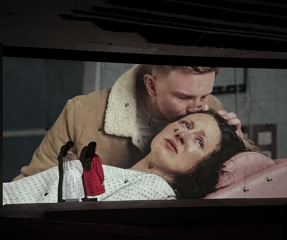 dwie aktorki na scenie przy ścianie, na której wyświetlane jest duże zdjęcie płaczącej kobiety i tulącego ją mężczyzny