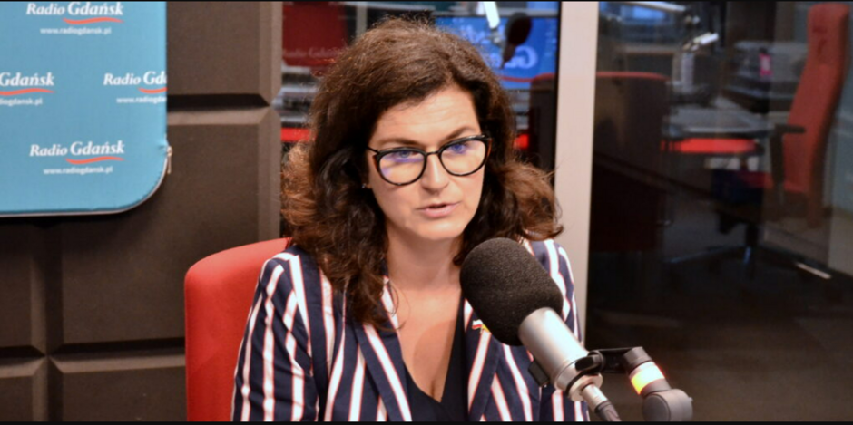 na zdjęciu kobieta w średnim wieku, szatynka w okularach, siedzi w studio radiowym, przed mikrofonem, zdjęcie do połowy, jest w granatowej marynarce w jasne paski