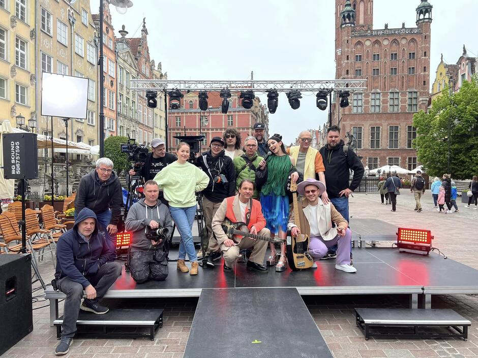 członkowie zespołu pozują z ludźmi do zbiorowego zdjęcia na małej scenie plenerowej ustawionej w Gdańsku na ulicy długiej