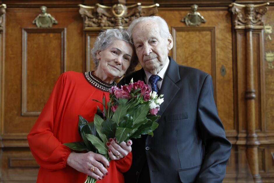 Dwoje staruszków elegancko ubranych, przytulonych do siebie głowami, patrzy w obiektyw. Seniorka trzyma bukiet czerwonych róż