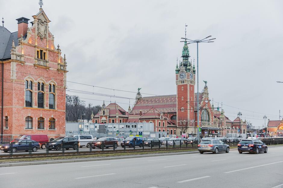 na zdjęciu fragment ruchliwej ulicy z samochodami, w tle zabytkowy budynek dworca, ceglany, zdobiony, po lewej inny zabytkowy ceglany budynek