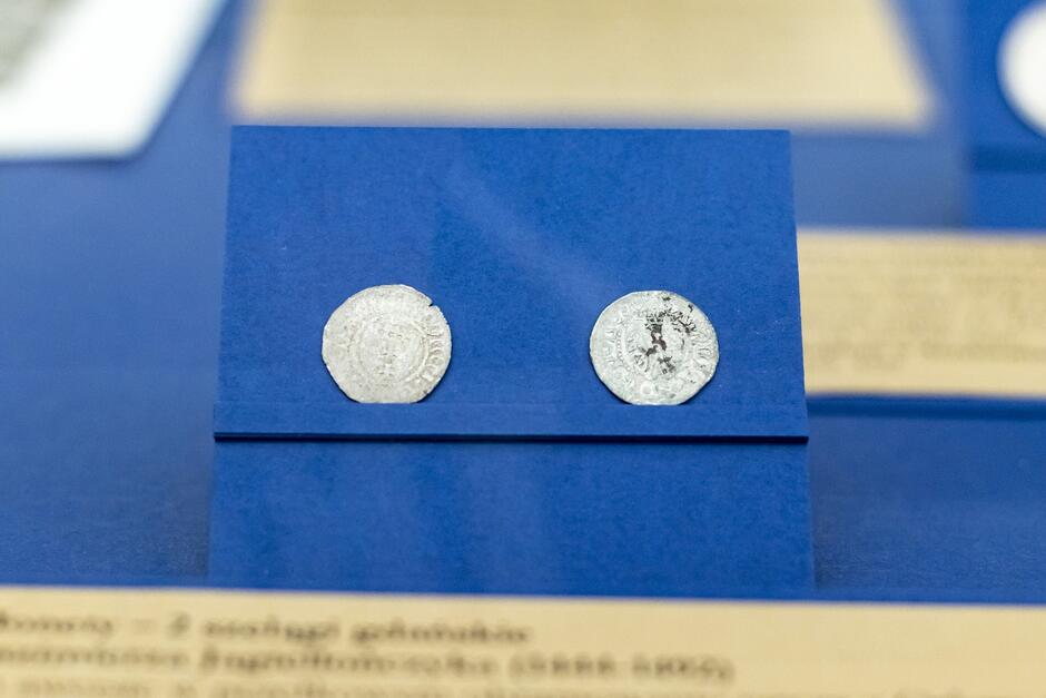 Na zdjęciu widoczne są dwa stare monety umieszczone na niebieskim tle. Monety są białe, być może srebrne, z wyraźnymi znakami korozji lub zabrudzenia, szczególnie jedna z nich ma ciemne plamy. Znaki i symbole na monetach są zużyte i trochę trudne do odczytania ze względu na ich stan. Monety są eksponowane w muzealny sposób, prawdopodobnie są częścią wystawy lub kolekcji. W tle, rozmyty przez niską głębię ostrości, widać fragment tekstu, który wydaje się być częścią opisu ekspozycji, jednak ze zdjęcia nie można dokładnie odczytać tekstu.