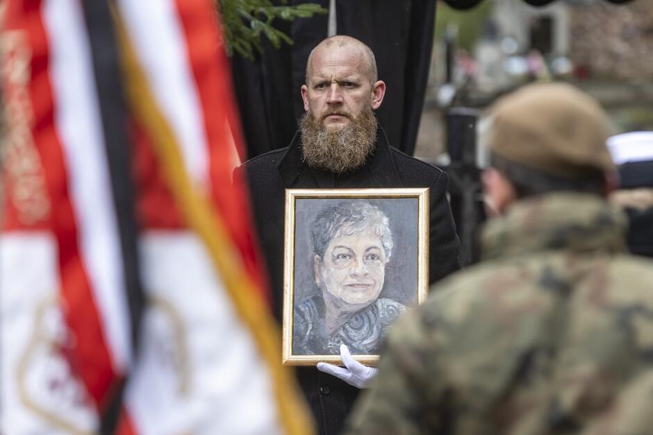 Łysy mężczyzna z długą brodą trzyma w dłoni, ubranej w białą rękawiczkę, portret kobiety w średnim wieku. Po lewej stornie zdjęcia widać nieostre biało-czerwone chorągwie, po prawej stojącego tyłem do zdjęcia żołnierza w stroju polowym.