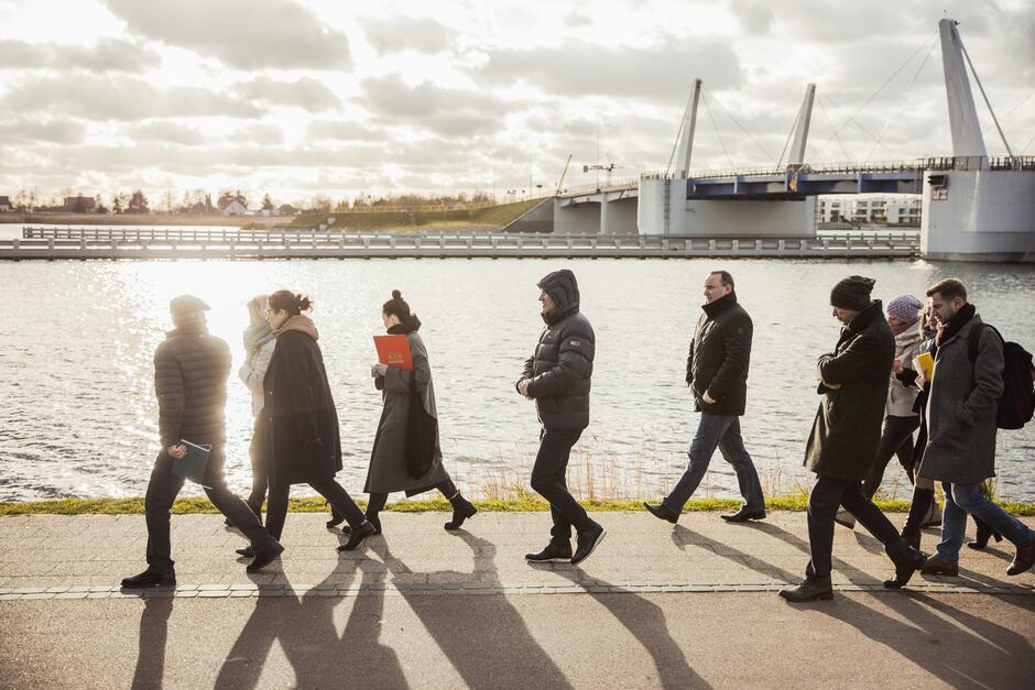 na zdjęciu kilka osób w zimowych ubraniach idzie, w tle most zwodzony i fragment rzeki
