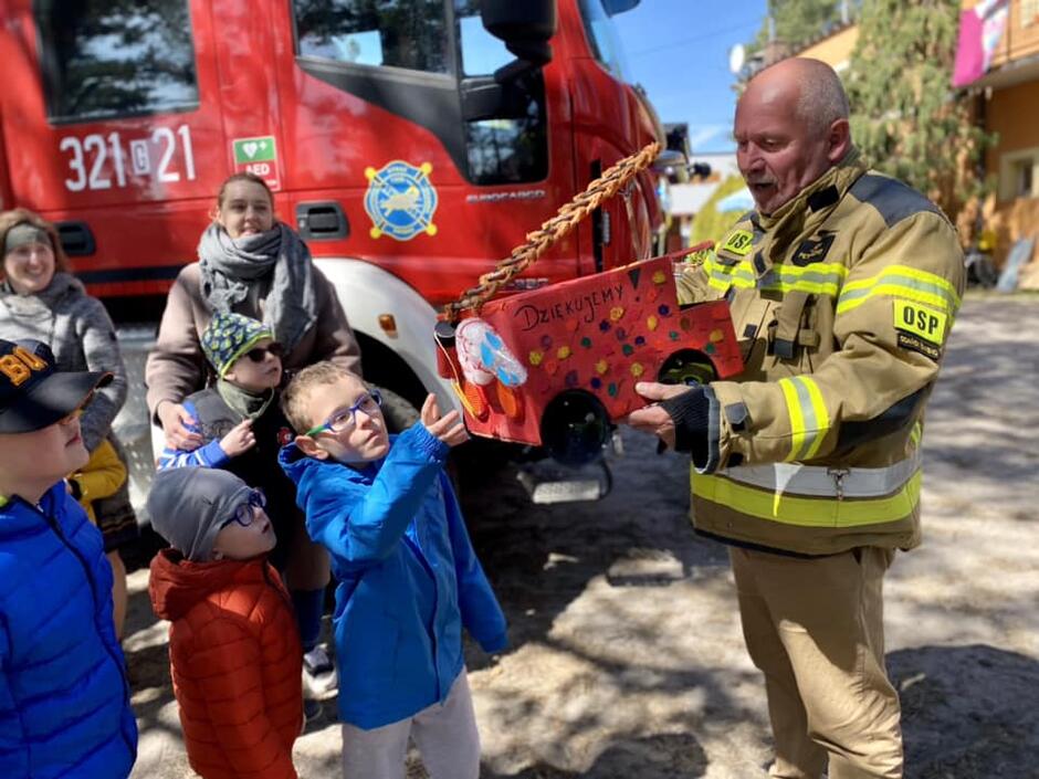 grupa dzieci i strażak, chłopiec przekazuje wykonany przez dzieci pojazd strażacki