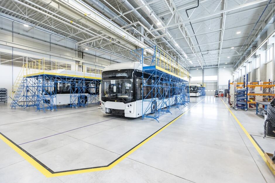 Na zdjęciu widać rozległą i bardzo jasną halę produkcyjną i stojące na środku dwa niekompletne autobusy w trakcie montażu.