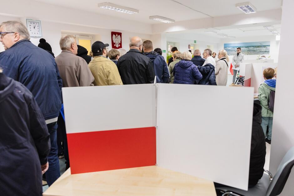 Na zdjęciu widzimy wnętrze lokalu wyborczego. Przestrzeń jest wypełniona ludźmi oczekującymi w kolejce, aby oddać swój głos. W tle, na ścianie, widnieje biało-czerwony herb Polski, co wskazuje, że to wydarzenie ma miejsce w Polsce. Przednia część zdjęcia pokazuje stolik wyborczy z przegrodą, która umożliwia tajne głosowanie. Lokal wydaje się być dobrze oświetlony i organizowany w sposób, który umożliwia sprawną obsługę wyborców.
