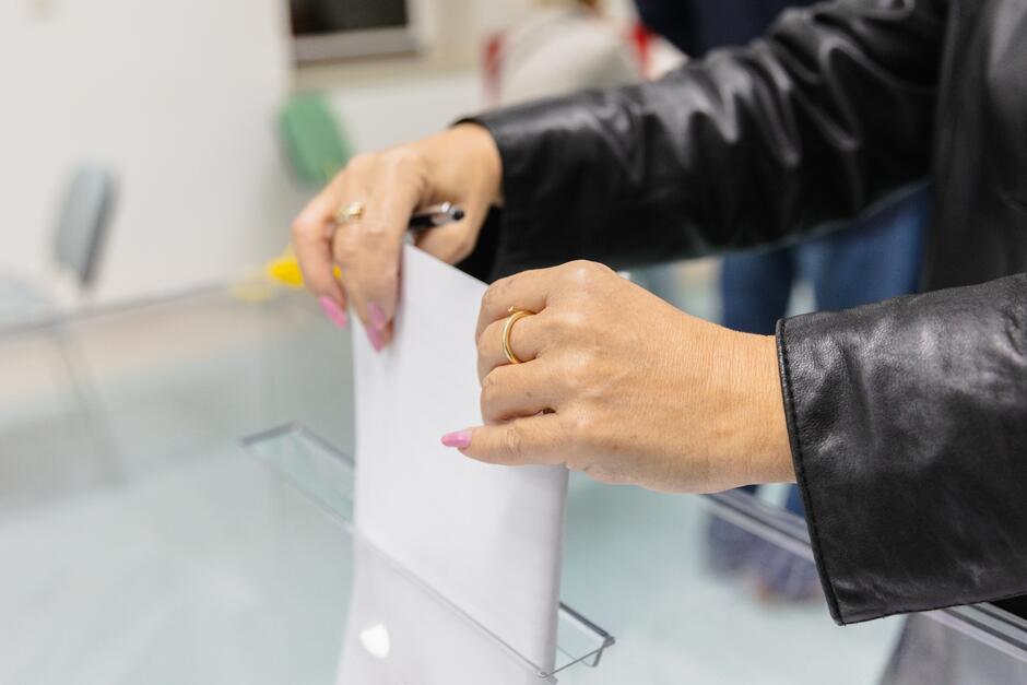 Zdjęcie pokazuje zbliżenie na ręce osoby wrzucającej kartę do głosowania do przezroczystej urny. Osoba ta ma na sobie czarną kurtkę i różowy lakier na paznokciach, a na palcu widoczny jest złoty pierścionek. To ujęcie podkreśla akt głosowania, będący istotnym elementem demokratycznego procesu wyborczego. Urna jest umieszczona na stoliku z przezroczystą osłoną, co może wskazywać na dodatkowe środki bezpieczeństwa lub higieny