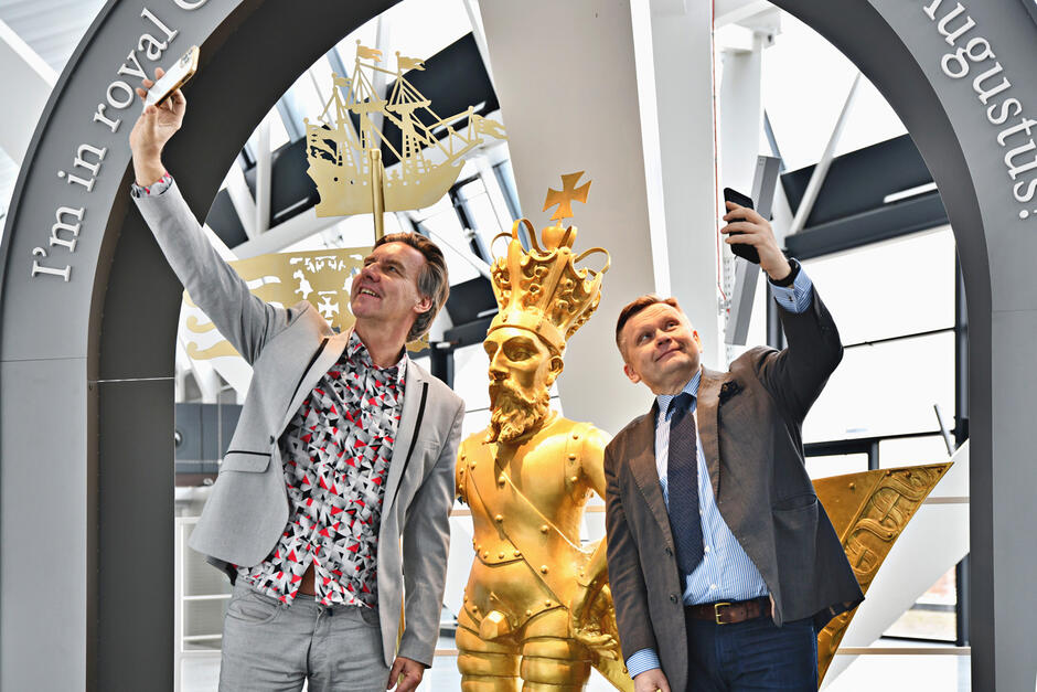 wymienieni mężczyźni w starszym wieku wykonują selfie, oraz eksponowana na postumencie ponad 2 metrowa złota rzeźba króla, 