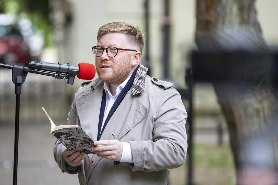 niewysoki rudy mężczyzna w okularach, elegancko ubrany, czyta do mikrofonu wiersz z trzymanej w dłoniach książki