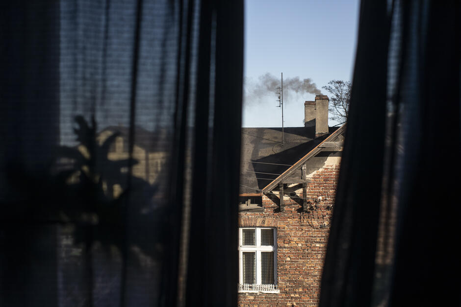 Zdjęcie zrobione z wnętrza mieszkania - jest to widok przez okno. Na zewnątrz znajduje się ściana sąsiedniego domu, z dachem i dymiącym kominem 