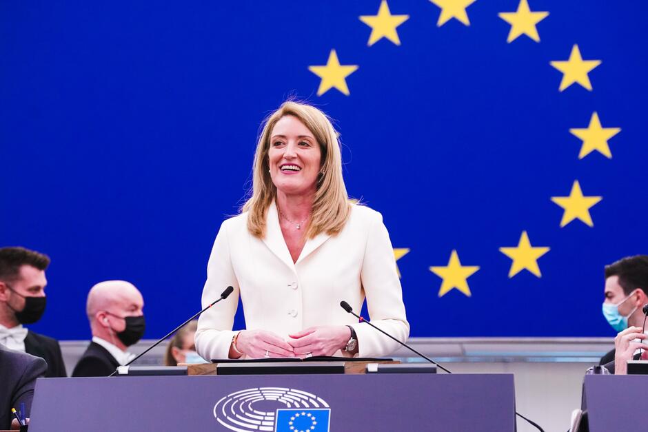 Młoda kobieta o długich jasnych włosach. Przemawia, stojąc na tle ściany z motywem flagi Unii Europejskiej