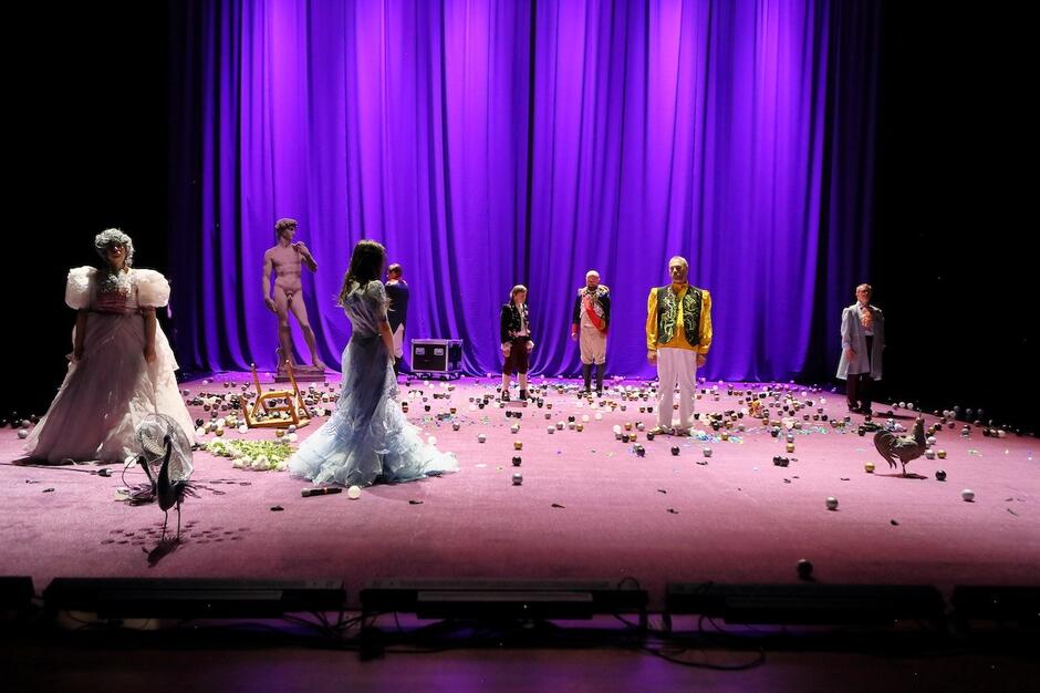 Na scenie oświetlonej na kolor różowy stoi kilku aktorów w powłóczystych kostiumach. Stoją w kręgu zwróceni do siebie twarzami.